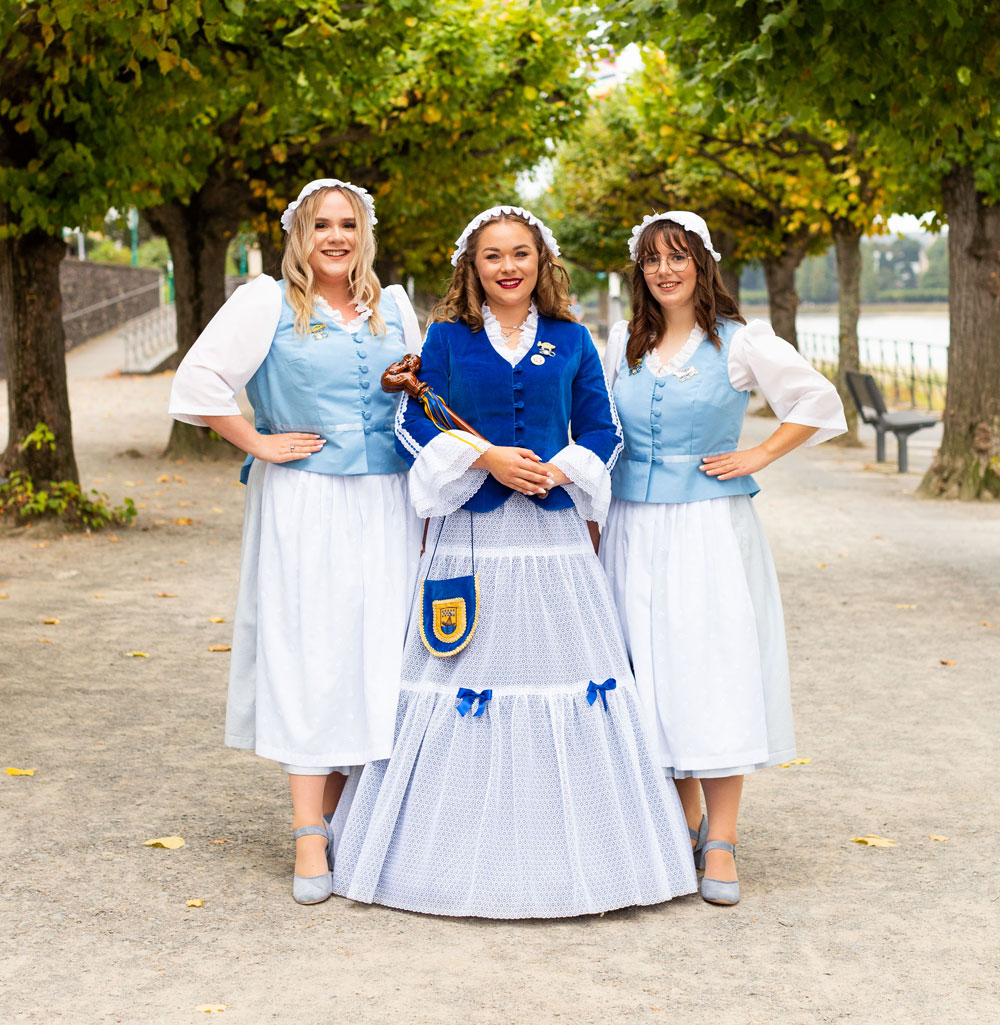 Drei Frauen in traditioneller Kleidung posieren lächelnd in einem von Bäumen gesäumten Parkweg.