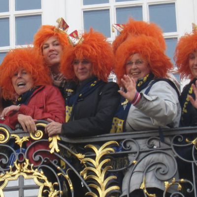 Fröhliche Frauen mit orangen Perücken winken vom dekorierten Balkon beim Sessionsauftakt der Beueler Weiberfastnacht.