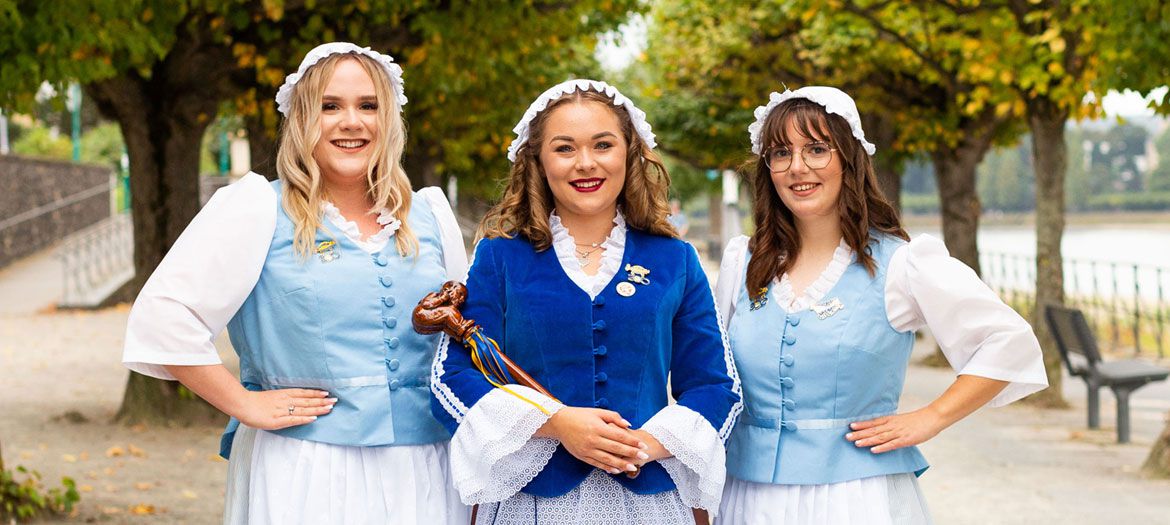 Drei Frauen in traditionellen Kostümen posieren lächelnd am Flussufer, passend zum Titel "Sabirina" der Weiberfastnacht.